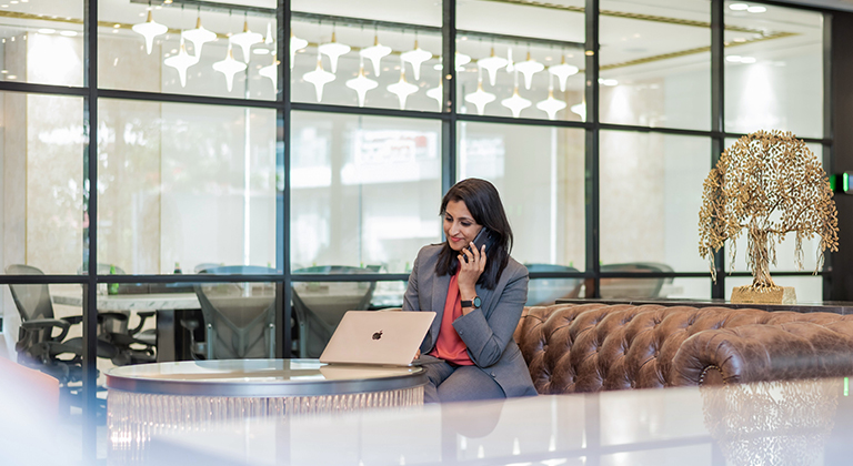 A businesswoman in a grey suit speaks on her phone while seated at a laptop in the Kaman Amaryllis centre lounge