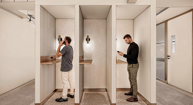 Two men using mobile phones while standing in phone nooks within a modern office