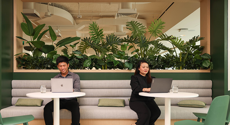Two colleagues engage with laptops at lounge tables surrounded by vibrant plants in The Exchange centre