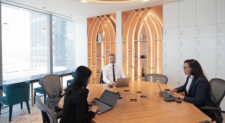 Three coworkers sit at a round conference table with laptops, discussing ideas in a meeting room with a city view