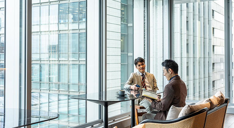 Two men seated at a table in a member lounge, engaged in conversation